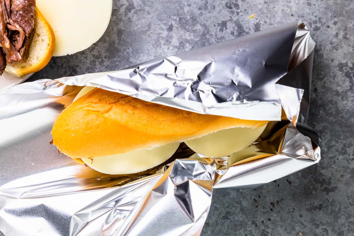 an assembled crockpot french dip sandwich being wrapped in aluminum foil.