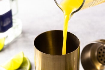 orange juice being poured into a metal cocktail shaker.