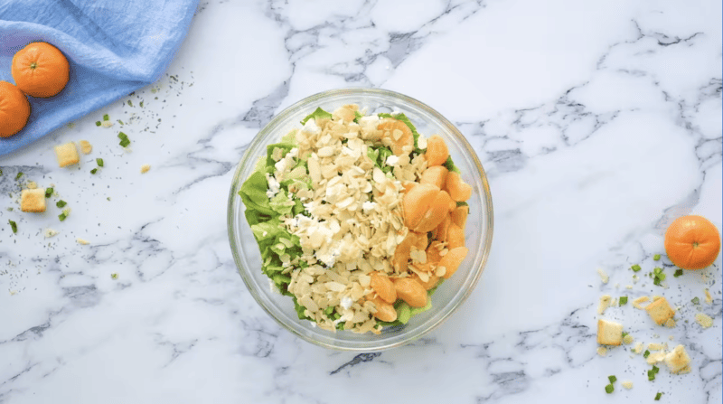 ingredients for mandarin orange salad in a glass bowl on a marble counter.