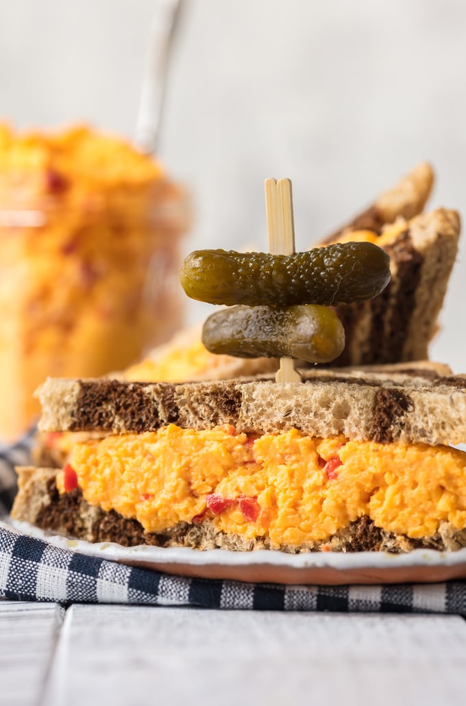 Want to learn How to Make Pimento Cheese? You've come to the right place! This Homemade Pimento Cheese recipe is bold and full of flavor. The classic Southern spread is perfect to serve at parties, and of course it makes an amazing Pimento Cheese Sandwich. With tasty Colby cheese and sweet Pimento peppers, this is the Best Pimento Cheese recipe out there! Pimento Cheeses Sandwich with bite-sized pickles on top