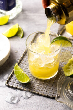 a cocktail shaker pouring a skinny margarita into a rocks glass filled with ice with a lime wedge on the rim.