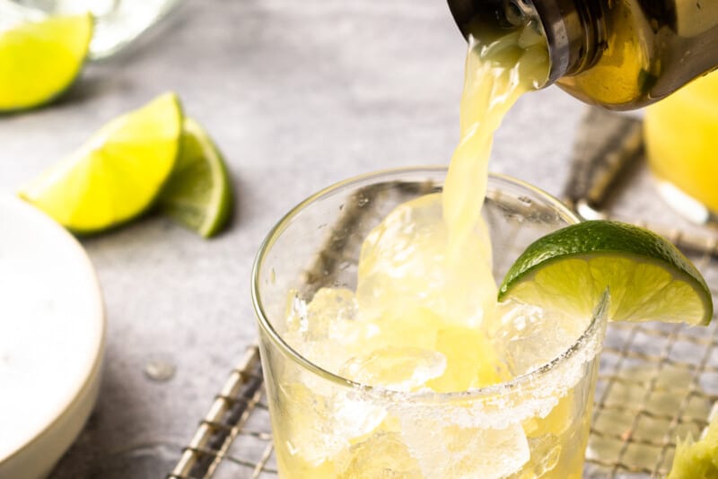 a cocktail shaker pouring a skinny margarita into a rocks glass filled with ice with a lime wedge on the rim.