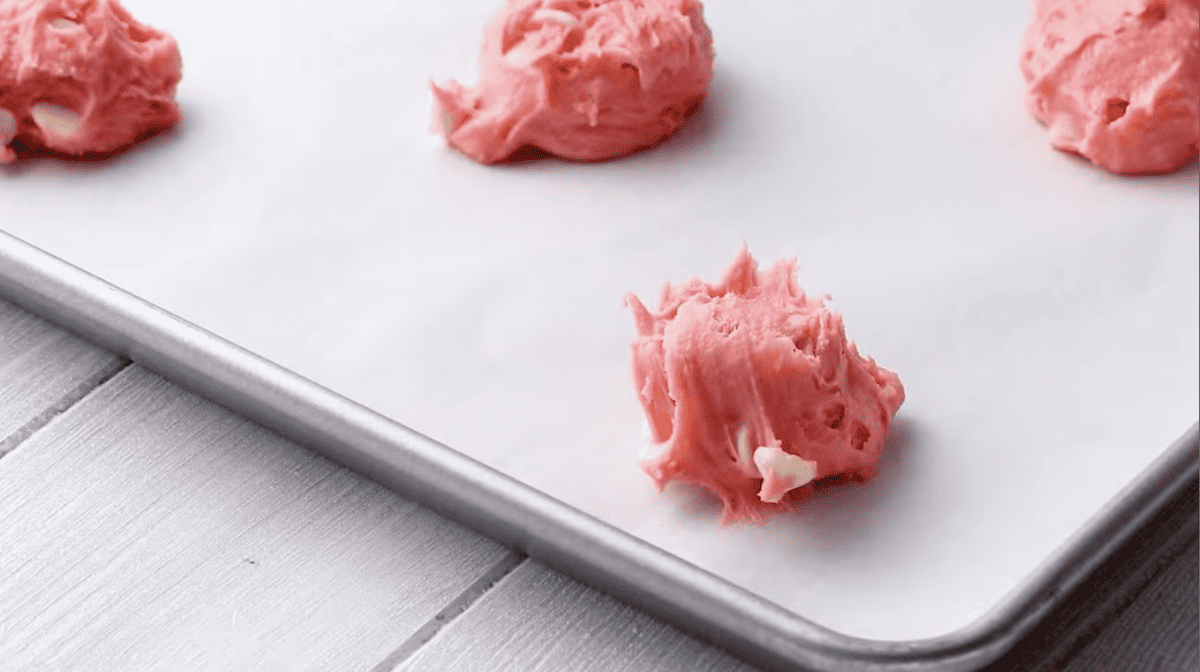 Pink cookie dough balls with white chocolate chips on a baking sheet.