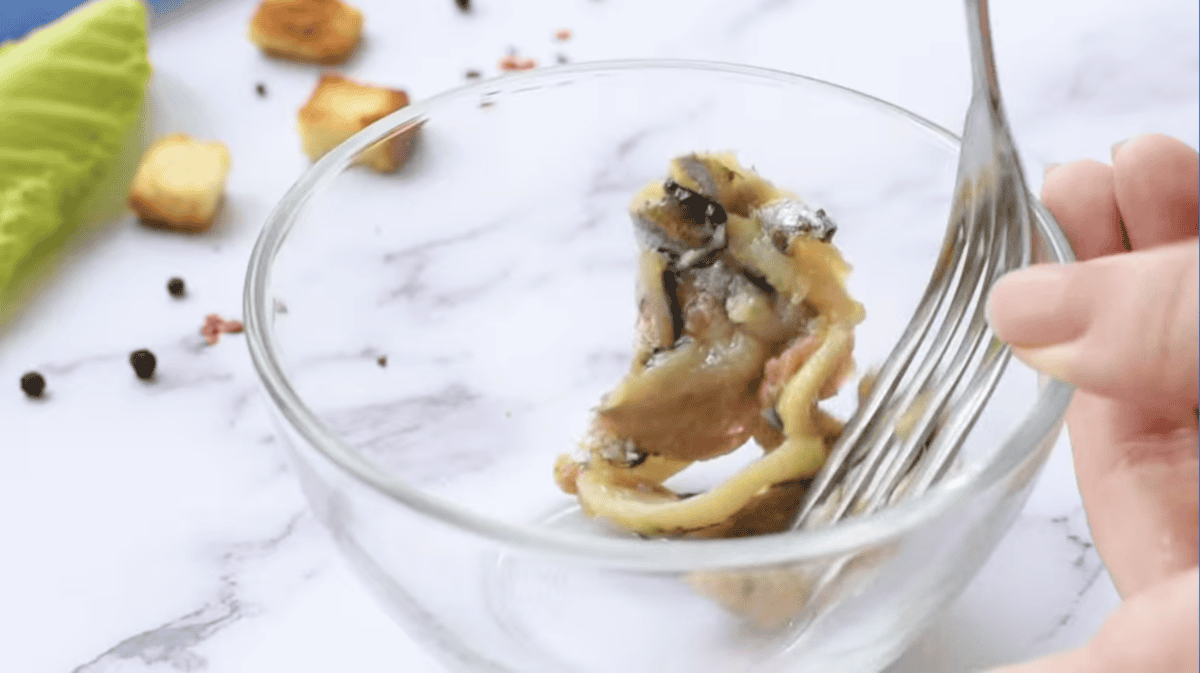 a fork mashing anchovies in a glass bowl.