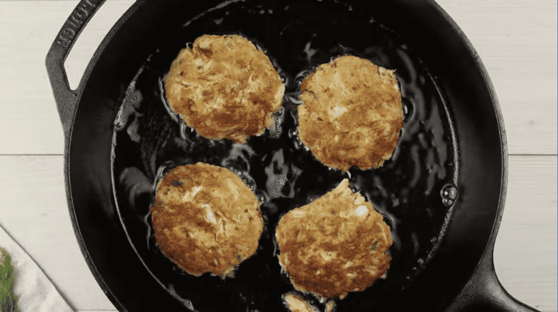 4 crab cakes cooking in a cast iron skillet.