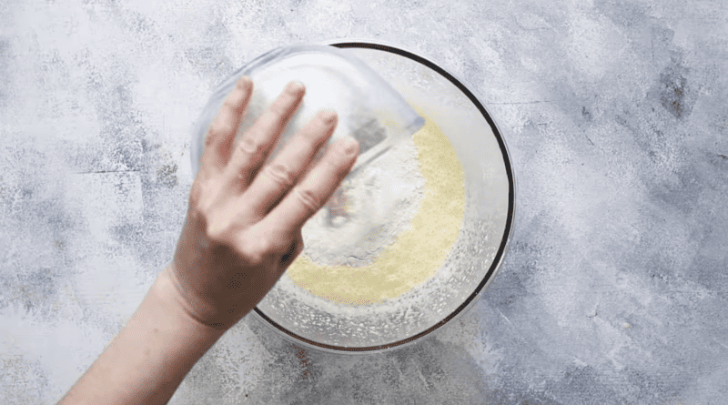 A hand is putting flour into a glass bowl to prepare blueberry pancakes.