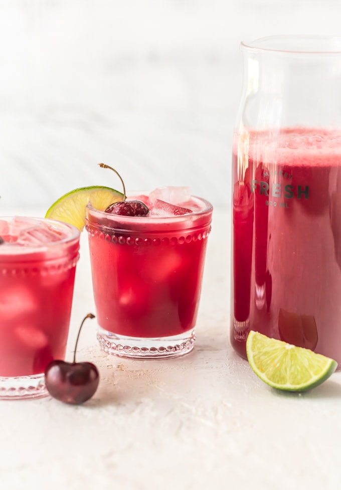 A pitcher of red cherry limeade next to two full glasses