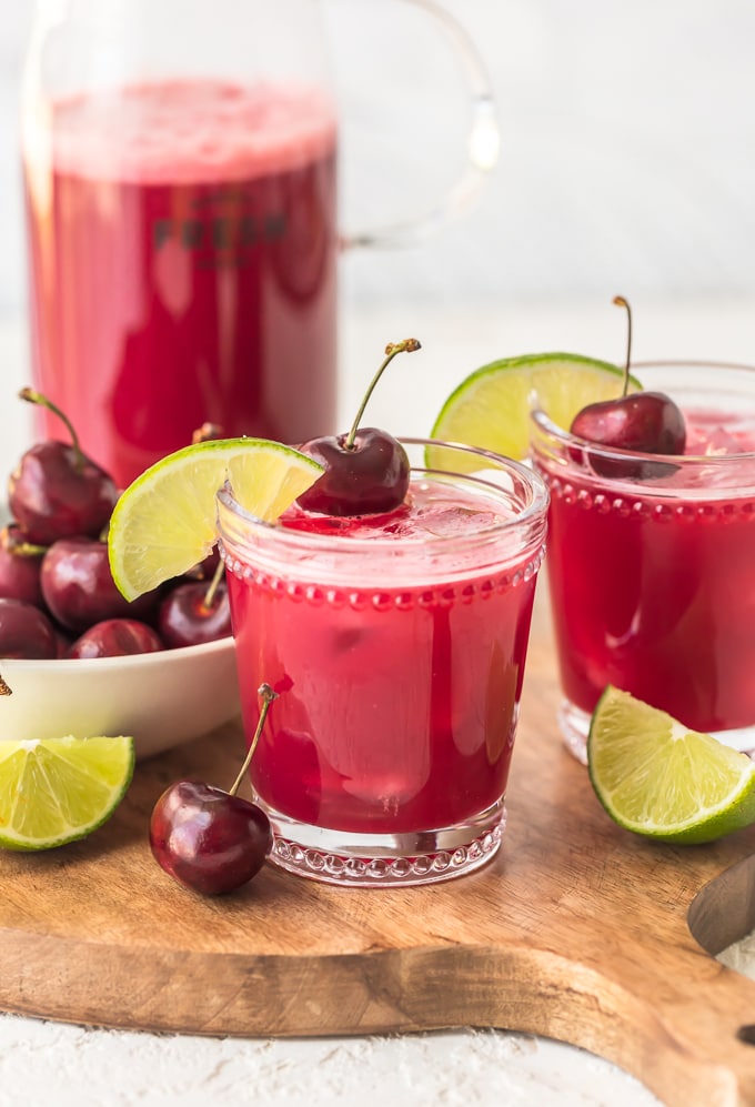 Two glasses of red cherry limeade next to a bowl of cherries