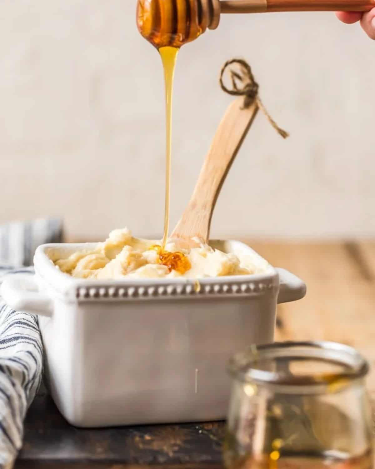 Honey butter in a white container with a wooden spoon and honey being poured on top.