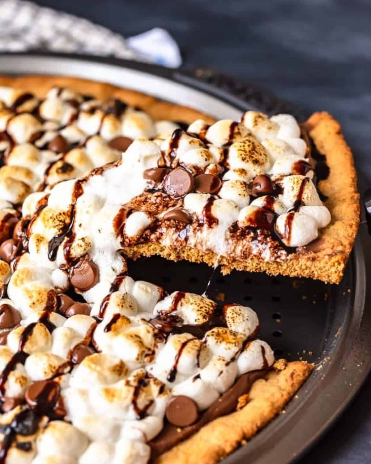 S'mores Dessert Pizza is a sweet, chocolaty dessert made just like a pizza! It's got the classic s'mores flavor of melted chocolate, graham crackers, and fluffy marshmallows, all melted together into the most amazing s'mores pizza. This easy dessert pizza recipe is fun to make and even more fun to eat! S'mores dessert pizza in a pizza pan with one slice being lifted.