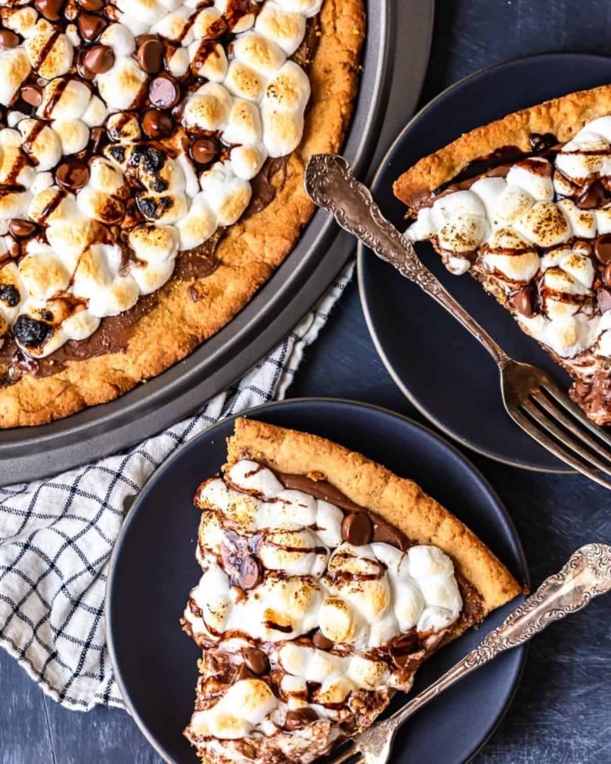 S'mores Dessert Pizza is a sweet, chocolaty dessert made just like a pizza! It's got the classic s'mores flavor of melted chocolate, graham crackers, and fluffy marshmallows, all melted together into the most amazing s'mores pizza. This easy dessert pizza recipe is fun to make and even more fun to eat! Slices of s'mores dessert pizza on plates with spoons.