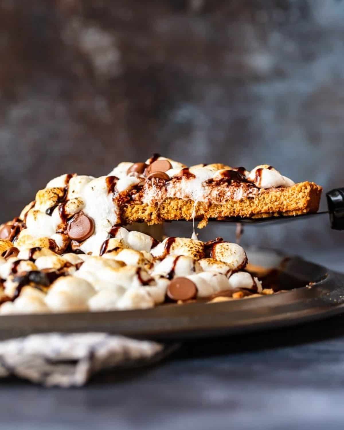 S'mores Dessert Pizza is a sweet, chocolaty dessert made just like a pizza! It's got the classic s'mores flavor of melted chocolate, graham crackers, and fluffy marshmallows, all melted together into the most amazing s'mores pizza. This easy dessert pizza recipe is fun to make and even more fun to eat! A slice of s'mores dessert pizza being lifted out of the pan.