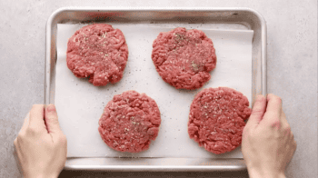 4 seasoned hamburger patties on a baking sheet.