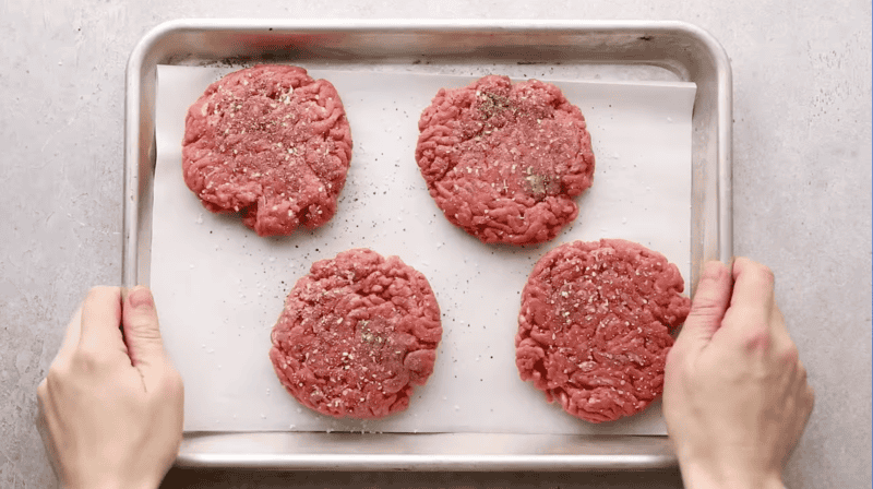 4 seasoned hamburger patties on a baking sheet.