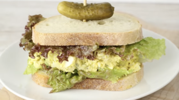 side view of an egg salad sandwich with lettuce and a pickle on a white plate.