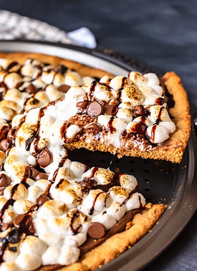 S'mores Dessert Pizza is a sweet, chocolaty dessert made just like a pizza! It's got the classic s'mores flavor of melted chocolate, graham crackers, and fluffy marshmallows, all melted together into the most amazing s'mores pizza. This easy dessert pizza recipe is fun to make and even more fun to eat! S'mores Dessert Pizza with slice cut out