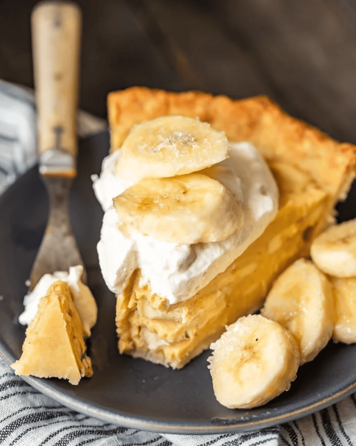 Banana Cream Pie is a creamy, delicious dessert filled with custard and fresh bananas. It's the perfect holiday recipe for Christmas, Easter, or Thanksgiving. This easy banana cream pie recipe is so rich and so tasty, everyone will love it! Slice of banana cream pie with slices of banana next to it.