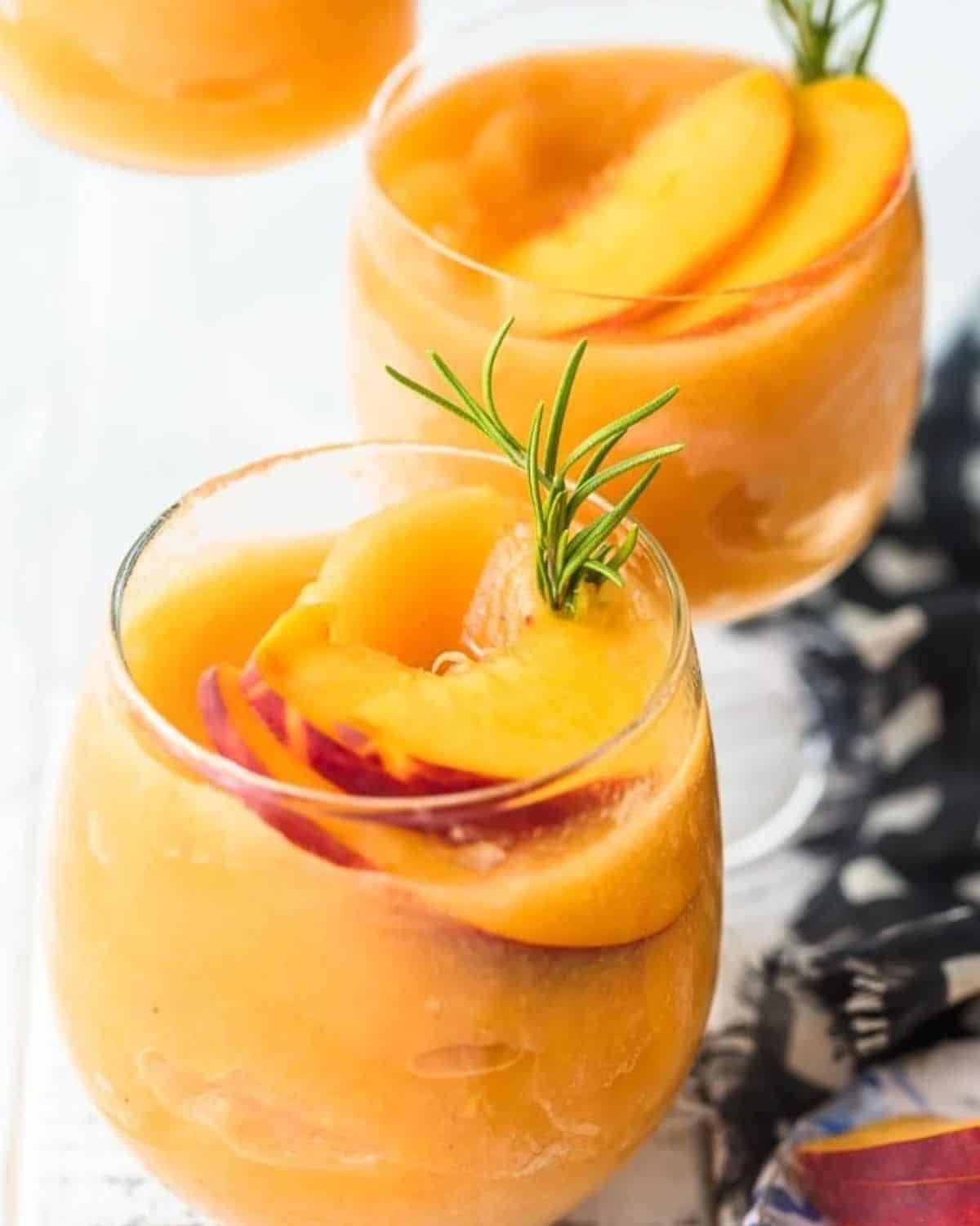 This Peach Frosé Recipe is a cocktail you definitely need for this hot Summer heat! All you need is frozen peaches, some Rosé wine, and a little honey. Frozen Rose is such a quick and easy fun cold drink to throw together for parties, friends and family, or just a quiet afternoon at home. We will show you how to make Frose and spice things up with this Peach Frose Recipe. So delicious, easy, fresh, and fun! Glass of peach frose garnished with fresh peaches and rosemary.