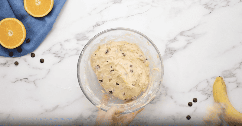 A person pouring chocolate chip banana bread batter into a bowl with oranges and bananas.