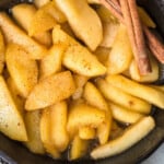 Fried sliced apples in a skillet with cinnamon sticks.