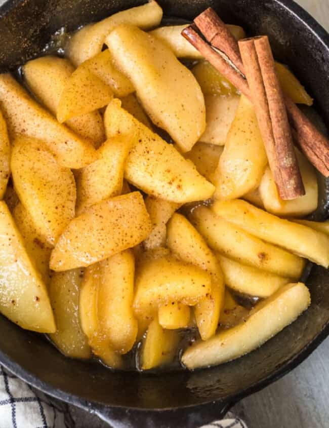 Fried sliced apples in a skillet with cinnamon sticks.