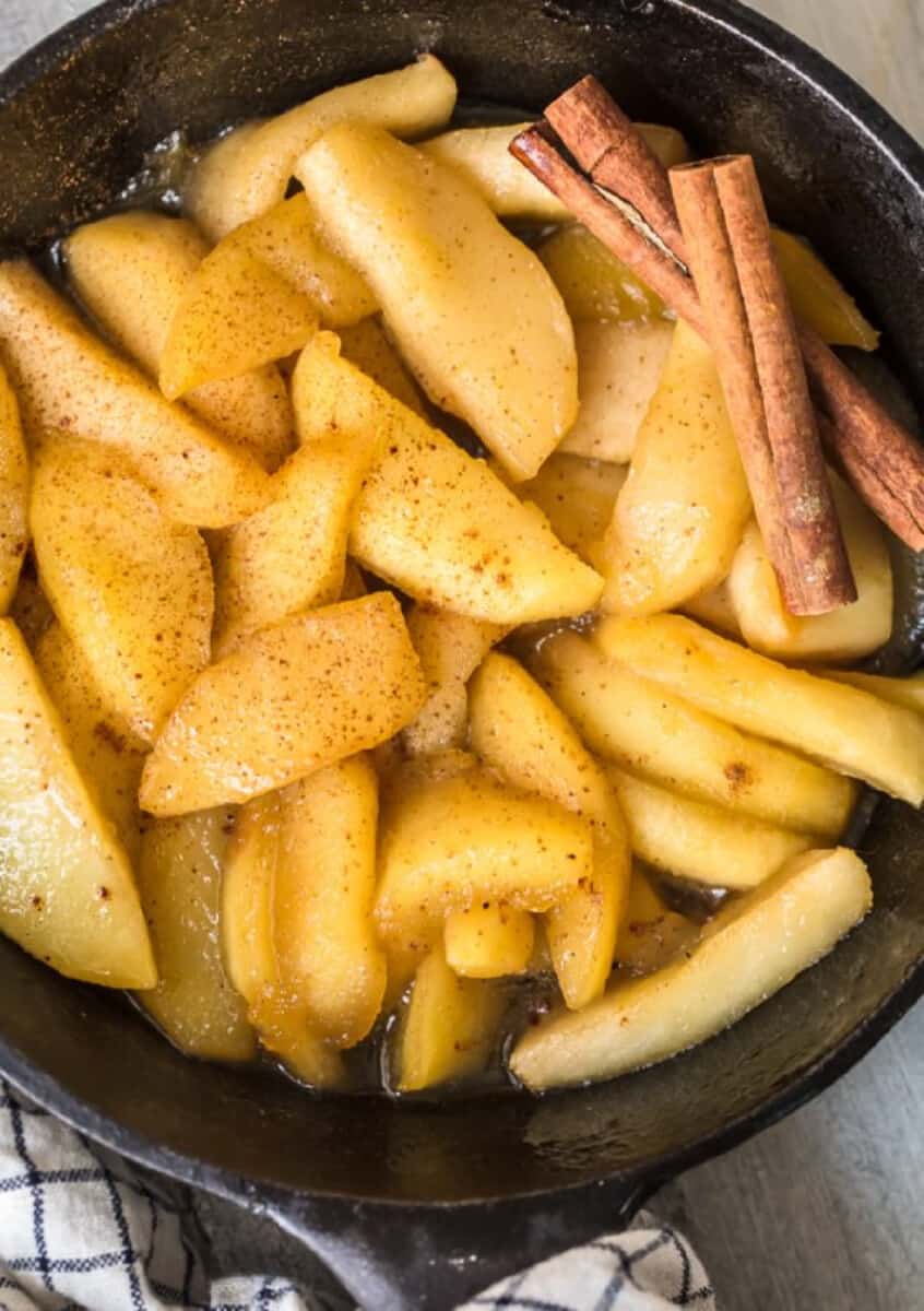 Fried sliced apples in a skillet with cinnamon sticks.