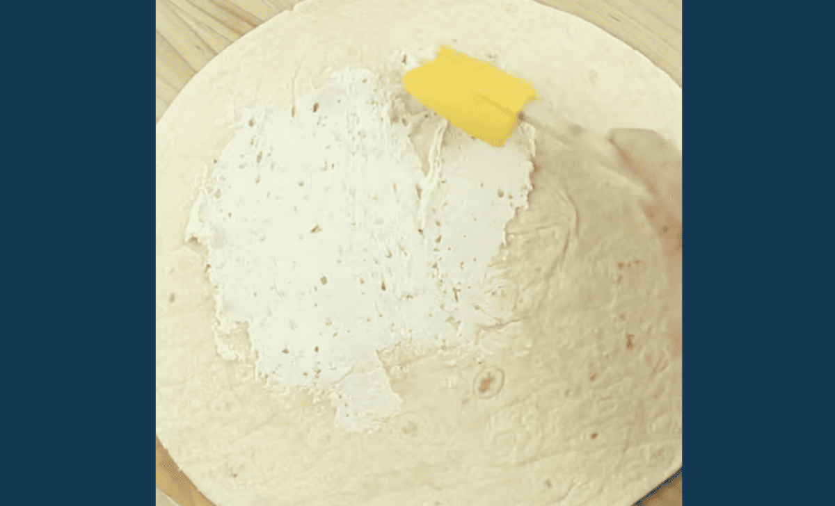 spreading boursin cheese over a flour tortilla with a yellow rubber spatula.