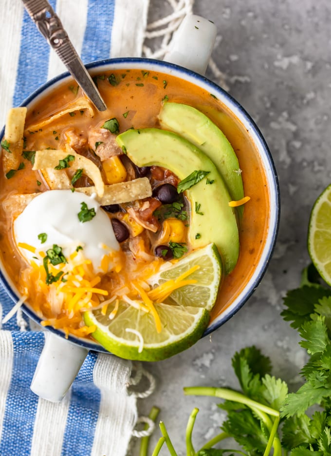 A bowl of creamy chicken tortilla soup topped with sour cream, cheese, limes, and avocado