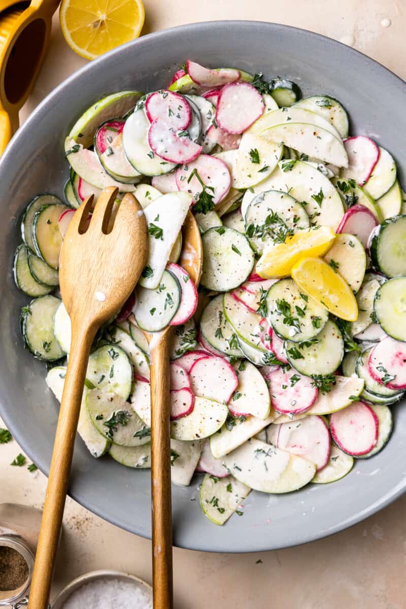 overhead view of creamy cucumber salad in a gray serving bowl with wooden salad serving utensils.