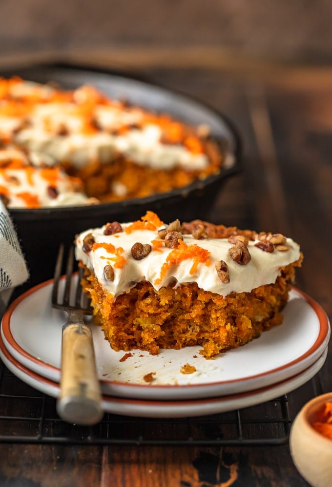 A slice of carrot cake on a plate, with a skillet carrot cake recipe in the bacground