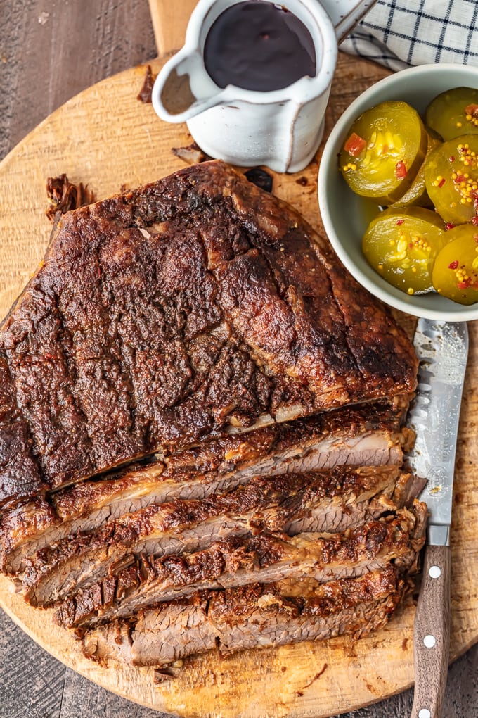overhead view of baked beef brisket on a cutting board, with a cup of BBQ sauce and a bowl of pickles set to the side