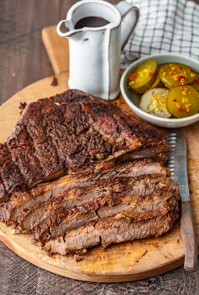 oven baked brisket on a cutting board, partially sliced, next to a bowl of pickles and BBQ sauce