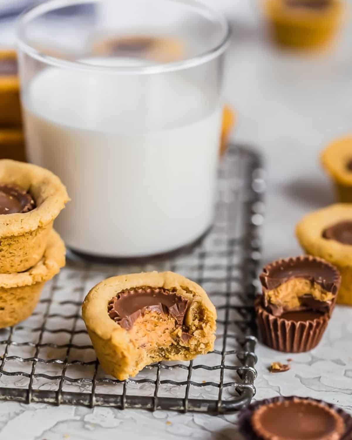 Peanut Butter Cup Cookies are the perfect little treat. Mini peanut butter cups are baked right into tart shaped cookies for a delicious bite-sized dessert. Everyone will eat these Reese's Cup Cookies right up because no one can resist chocolate peanut butter cookies! Peanut butter cup cookie on a cooling rack with a bite taken out of it and a glass of milk behind it.