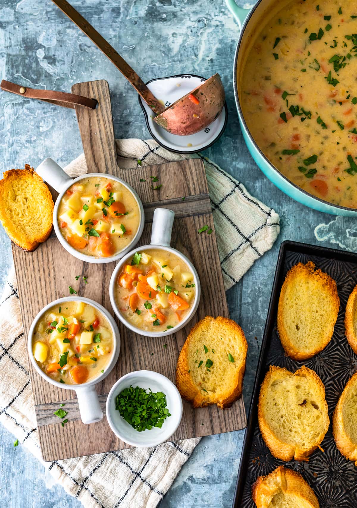 three bowls of soup on a wood cutting board, next to a pot of soup and a baking tray filled with toasted slices of bread