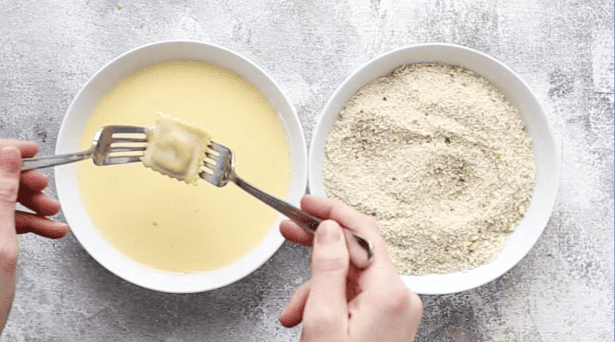 two forks holding a wet ravioli above an egg and milk mixture in a bowl next to a bowl of breadcrumbs.