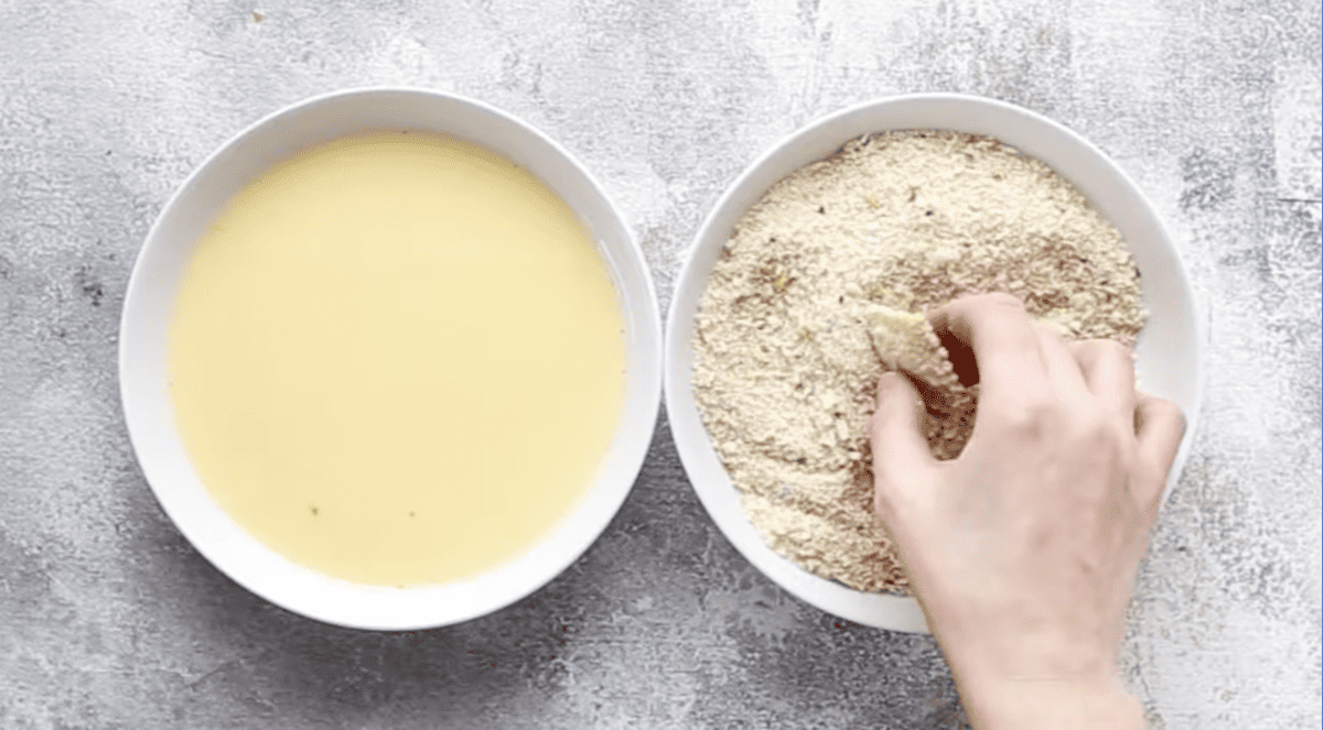 a hand dipping a ravioli in a bowl of breadcrumbs next to a bowl of egg and milk.