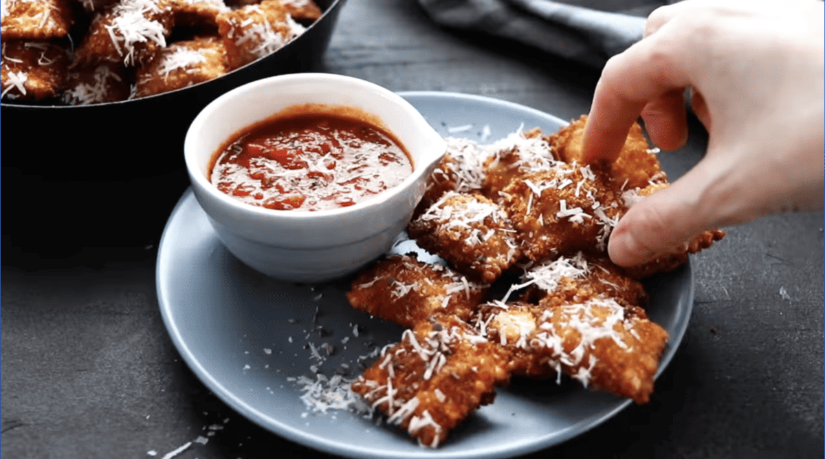 a hand reaching for a toasted ravioli coated in parmesan cheese on a plate.