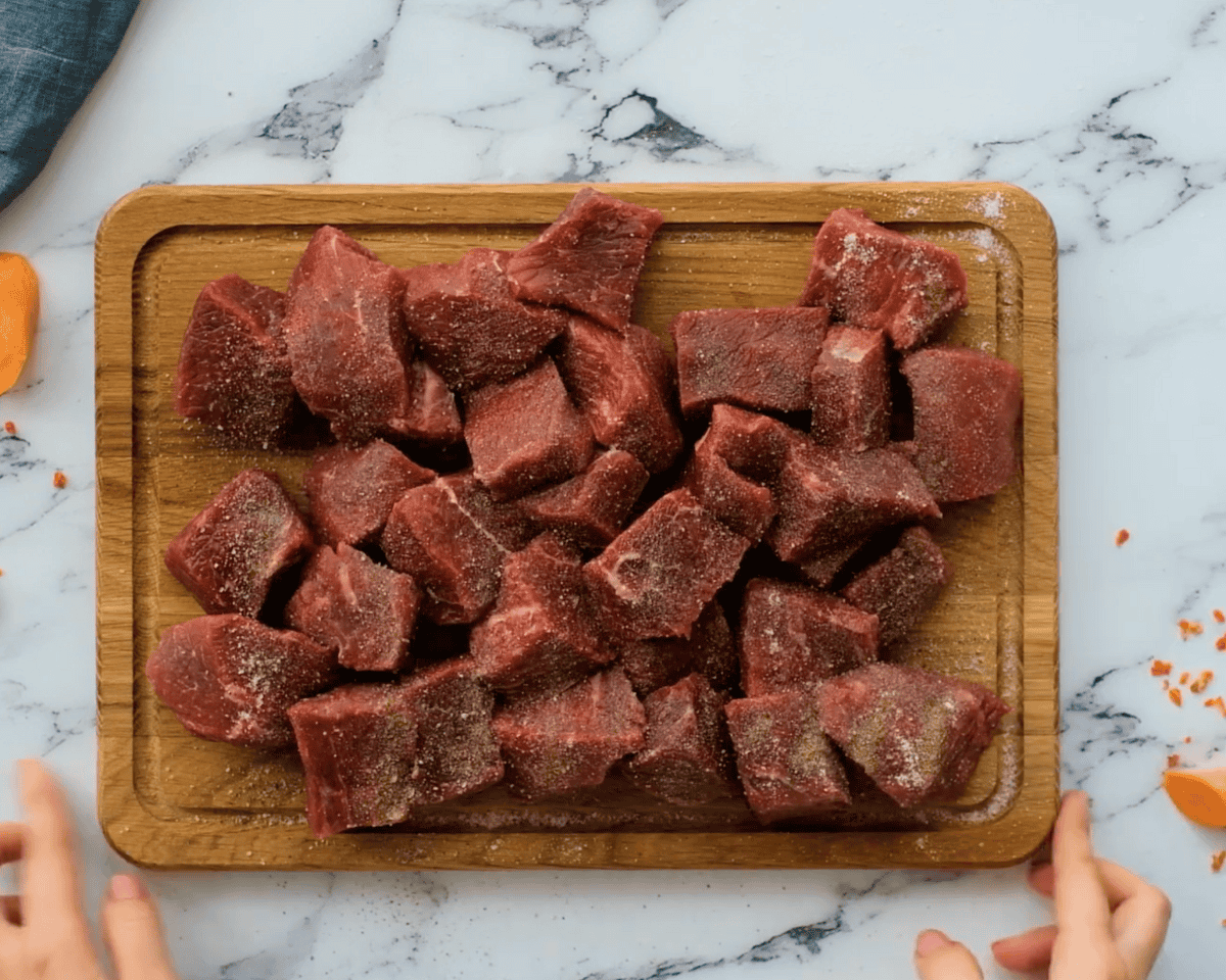 A top-down view of raw beef cubes sprinkled with salt and pepper on a wooden cutting board, ready for searing.