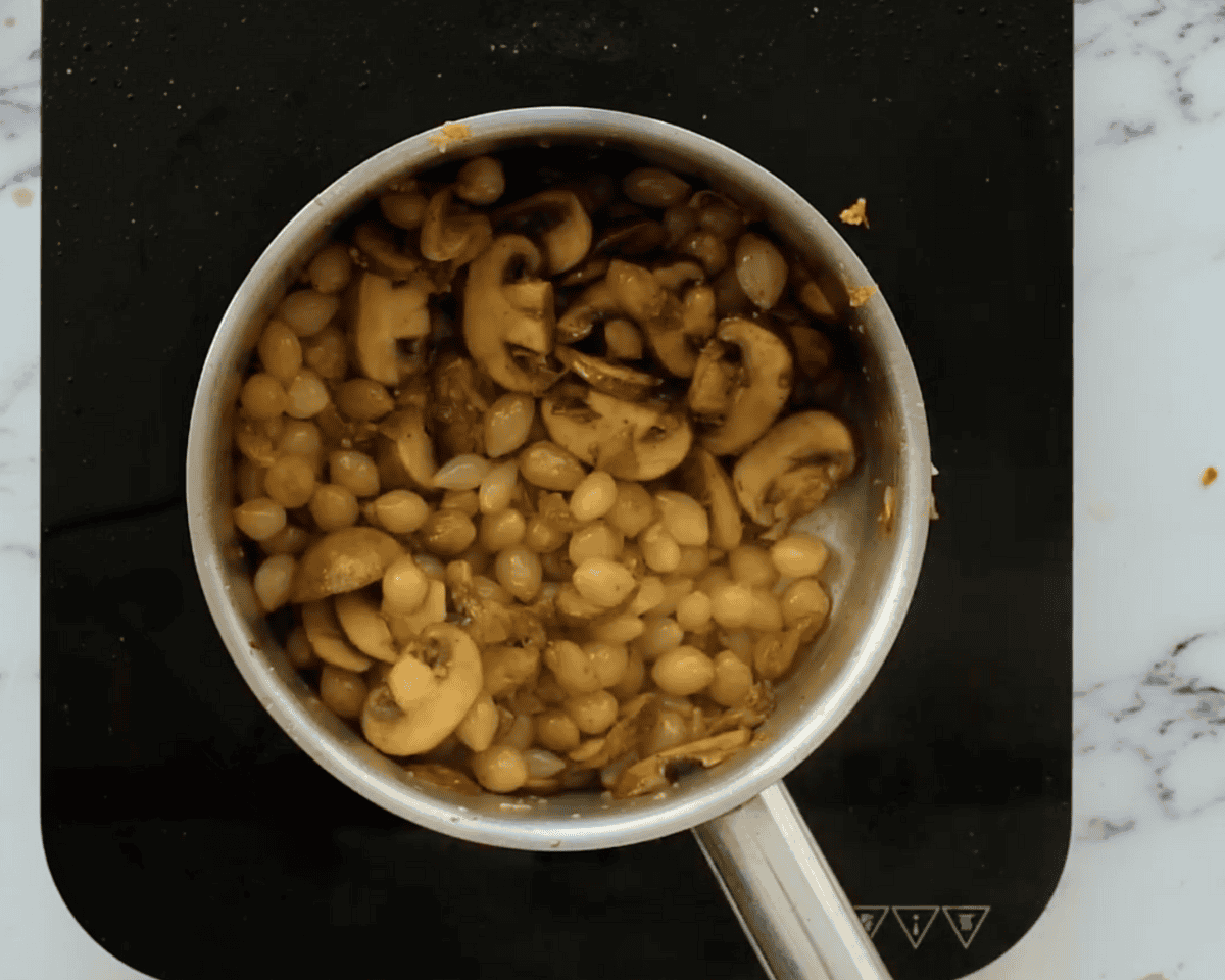 A saucepan filled with golden-brown sautéed mushrooms and pearl onions cooked in butter.