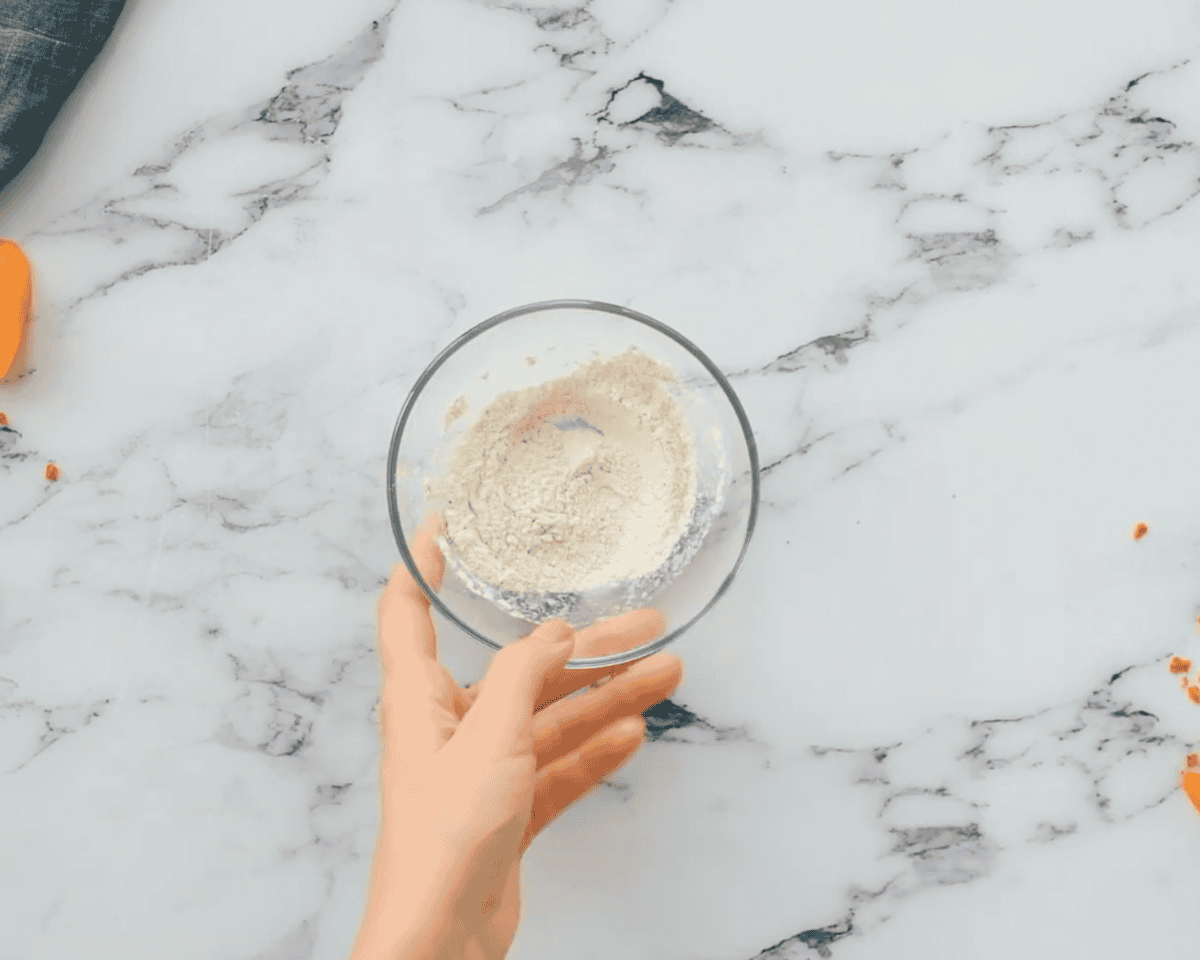 A hand holding a small glass bowl filled with a seasoned flour mixture on a marble countertop.
