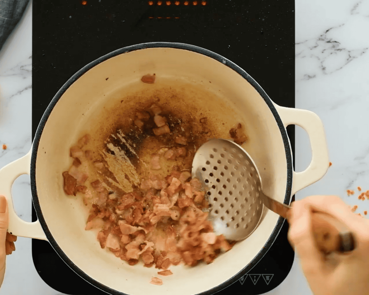 Bacon pieces sizzling and browning in a cream-colored Dutch oven as a slotted spoon stirs them.