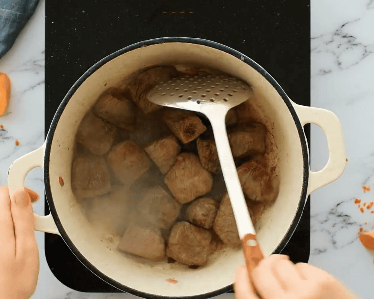 Browned beef cubes cooking in a Dutch oven, with a slotted spoon turning the pieces.