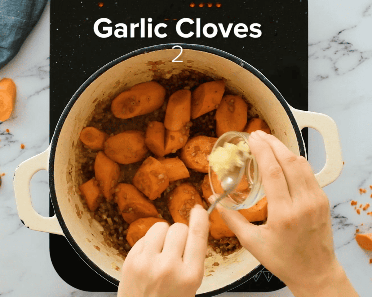 A spoonful of minced garlic being added to sautéed carrots and onions in a Dutch oven.