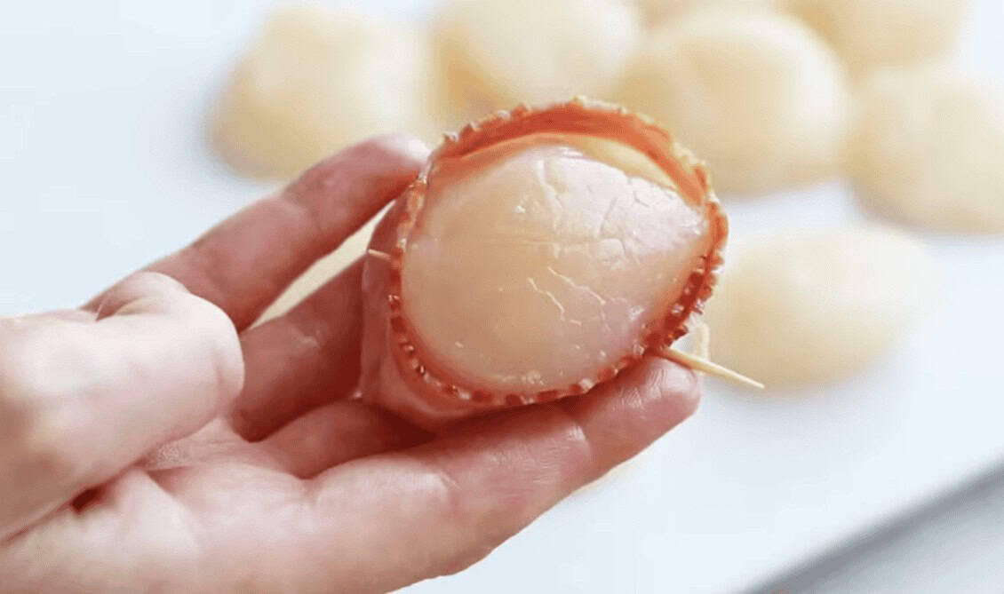 A hand holding a scallop wrapped in bacon.