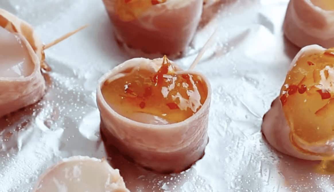 Raw bacon wrapped scallops on a foil lined baking sheet topped with red pepper jelly.