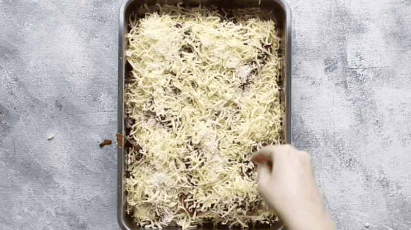 A person putting cheese into a lasagna baking pan.