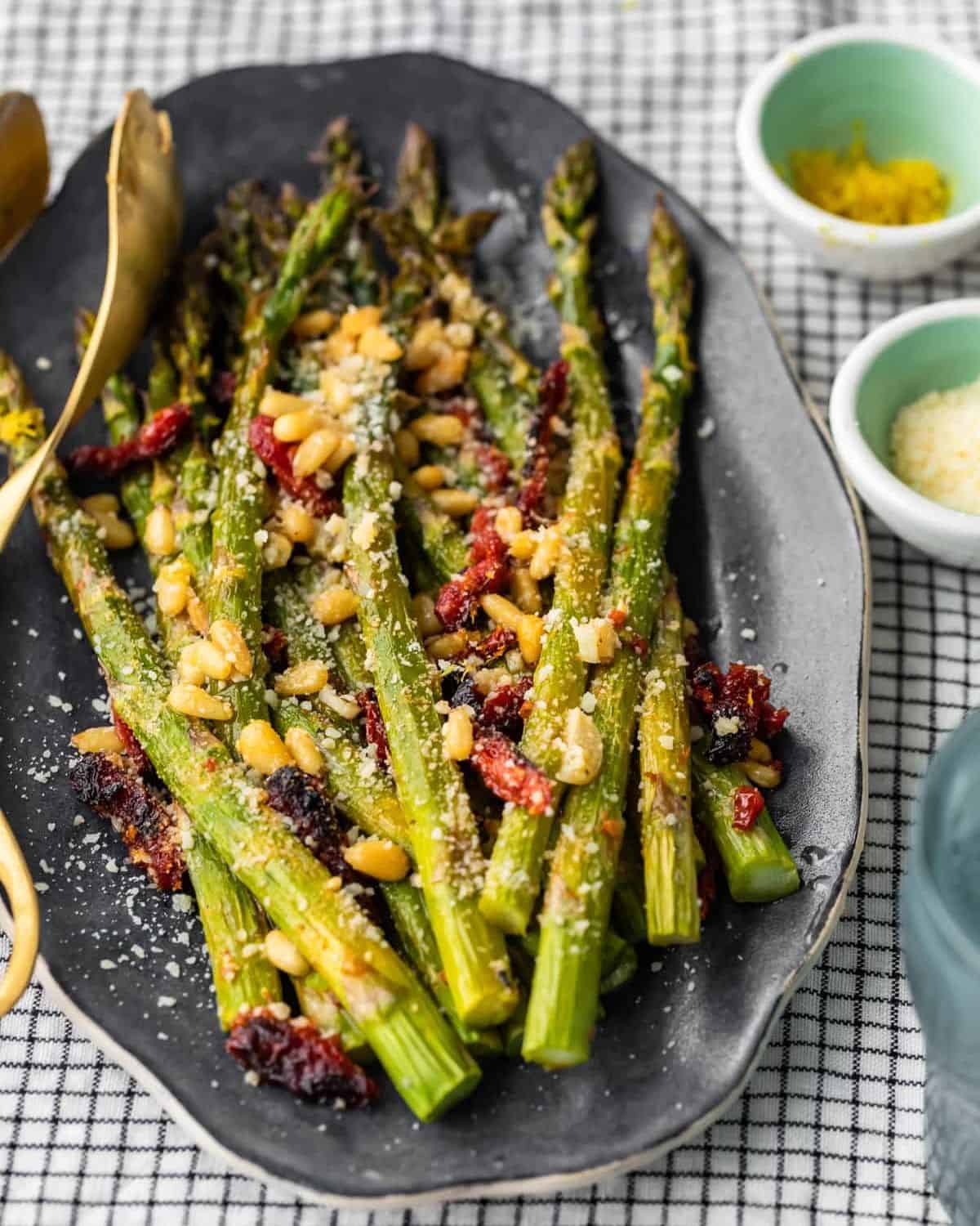 Oven Roasted Asparagus with sun dried tomatoes, pine nuts, garlic, and more is a delicious Mediterranean-inspired side dish fit for any occasion. This easy roasted asparagus recipe is quick to make yet filled with flavor! Add this asparagus side dish to your holiday meals, or just to your weeknight dinners. Either way, it's sure to be a hit! Oven roasted asparagus with sundried tomatoes, pine nuts, and Parmesan cheese on a platter.