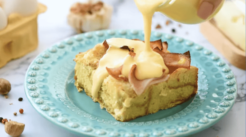 pouring hollandaise sauce over a slice of eggs benedict casserole on a blue plate.