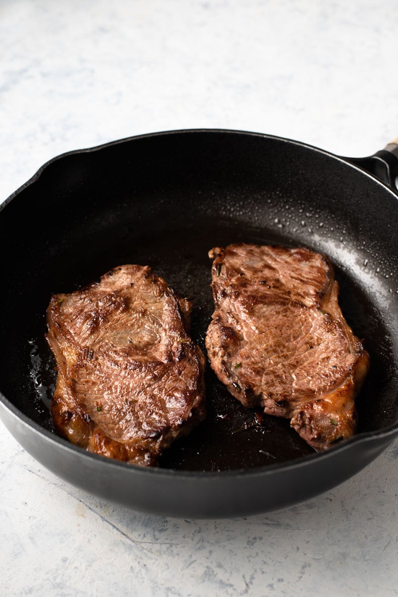 2 steaks cooking in a cast iron pan.