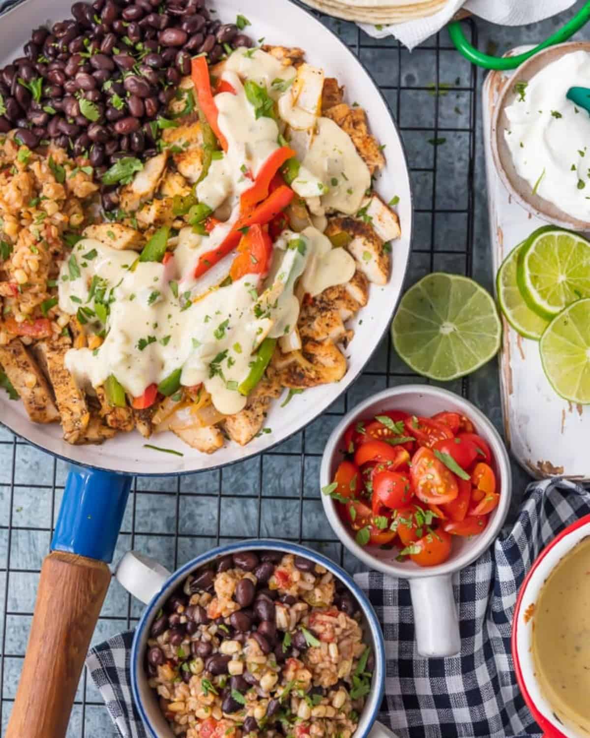 This easy Chicken Fajitas recipe has quickly become a favorite around here. AQueso Smothered Chicken Fajita Recipe with all the best ingredients! Queso smothered chicken in a skillet with bowls of chopped tomatoes, rice and beans and cheese sauce next to it.
